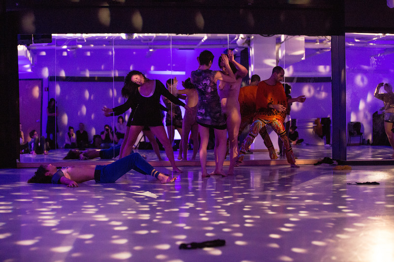 A mass of performers dancing against a mirror under a disco ball and purple lights